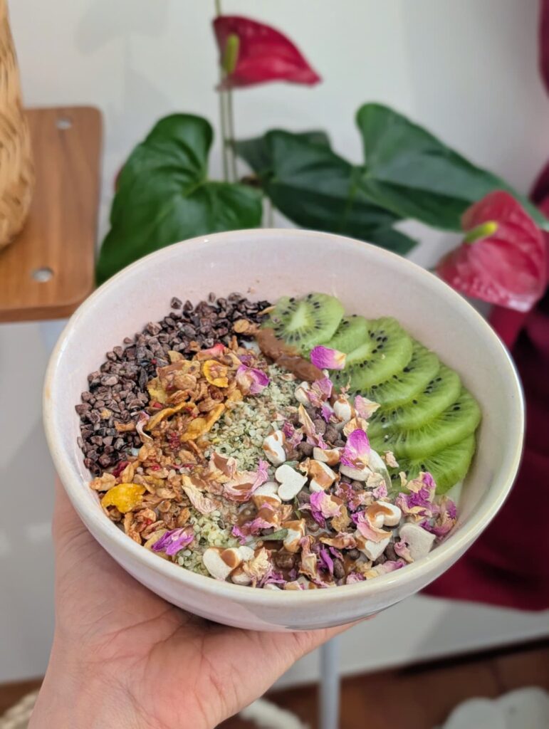 Christina Dengg hält eine Frühstücksbowl. Darin sieht man Kiwi-Scheiben, Cornflakes, Schokoflocken, Blütenblätter und kleine Herzen.